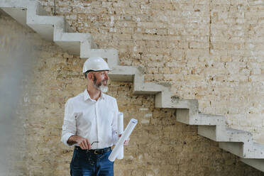 Happy architect wearing hardhat standing with blueprints at site - YTF01452