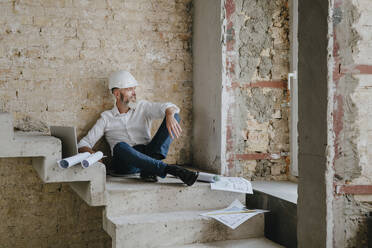 Lächelnder Architekt mit Laptop und Bauplänen auf einer Treppe vor Ort - YTF01457