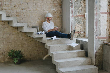 Architect holding coffee cup and sitting with blueprints on steps - YTF01458