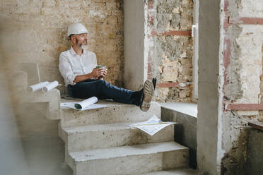 Nachdenklicher Architekt, der eine Kaffeetasse hält und mit Bauplänen auf einer Treppe sitzt - YTF01459