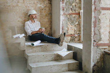 Thoughtful architect holding coffee cup and sitting with blueprints on steps - YTF01459