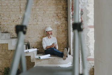Contemplative architect holding coffee cup and sitting with blueprints on steps - YTF01460