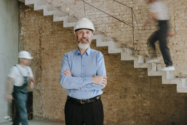 Happy architect standing with arms crossed at construction site - YTF01465