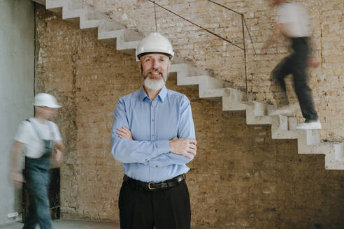 Happy architect standing with arms crossed at construction site - YTF01465