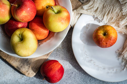 From below of collection of ripe, colorful apples presented on a rustic wooden board, with a white plate and draped fabric creating a serene kitchen setting - ADSF49517