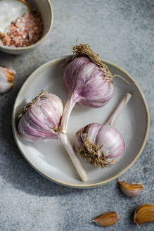 Top view of shot of raw garlic bulbs with purple streaks set against a grey background alongside a dish with coarse salt - ADSF49519