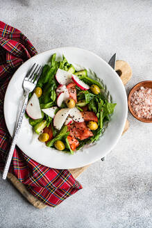 A vegetable salad made with fresh organic spinach leaves, sliced tomatoes, radishes, and olives presented in a white bowl beside a plaid cloth, wooden cutting board, and a bowl of pink salt - ADSF49525