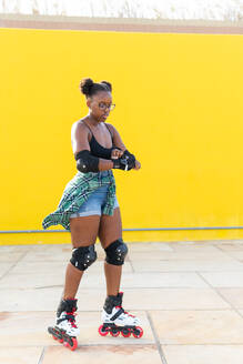 Full length of young African American woman fastening hand gear while rollerskating at skate park against yellow wall - ADSF49552