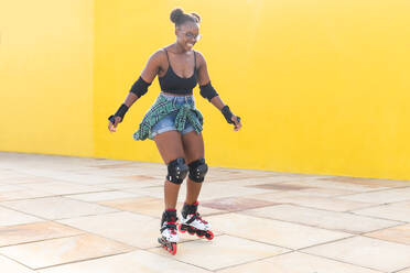 Full body of young woman smiling while rollerskating at skate park along yellow wall - ADSF49553