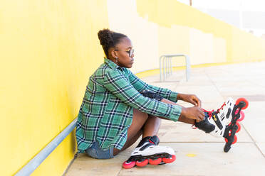 Side view full length of young woman wearing roller skate shoe while sitting by yellow wall at skate park - ADSF49554