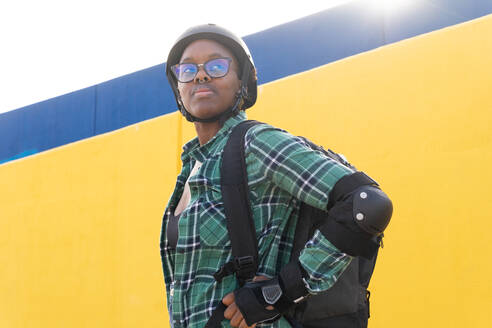 Thoughtful young woman in helmet and eyeglasses looking away while standing at skate park during sunny day - ADSF49555