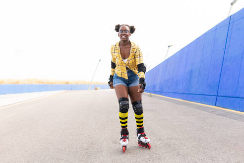 Full body portrait of happy African American woman rollerskating on rink at skate park against cloudless sky with lots of copy space - ADSF49559