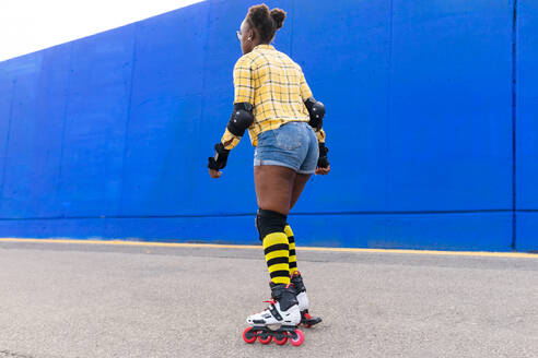 Full body back view of young African American woman rollerskating on rink at skate park along blue wall - ADSF49560