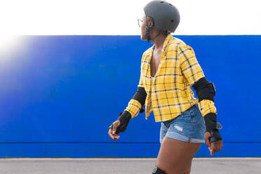 Side view of young woman in protective gear looking away while roller skating at skate park during sunny day - ADSF49563