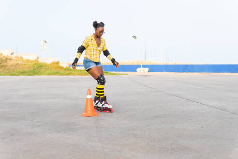 Full body of young African American woman rollerskating by cone while practicing on rink at skate park - ADSF49565