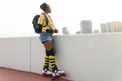 Thoughtful young African American woman in roller skates carrying backpack while standing at skate park - ADSF49566