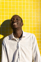 Cheerful black young man laughing against a vibrant yellow tiled wall, with the sun casting a shadow beside him - ADSF49568