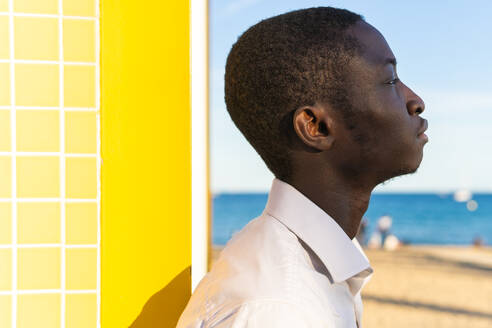 Side view young African American man gazes towards the ocean against a bright yellow backdrop and a tiled wall - ADSF49569