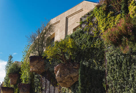 From below of beige building adorned with a lush vertical garden featuring various green plants, shrubs, and rocks, set against a clear blue sky - ADSF49578