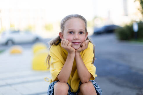 Joyful blonde girl wearing a yellow top seated on a curb with her chin rested on her hands smiles brightly against an urban backdrop looking at camera - ADSF49580