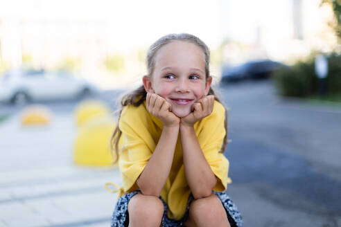 Joyful blonde girl wearing a yellow top seated on a curb with her chin rested on her hands smiles brightly against an urban backdrop looking away - ADSF49581