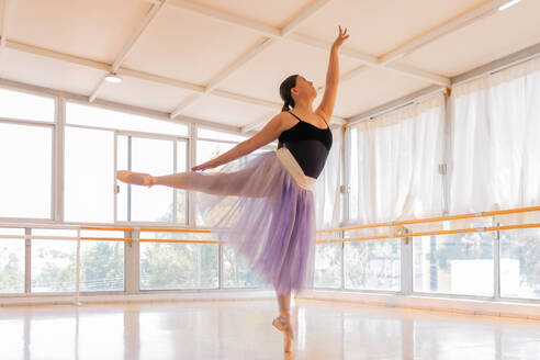 Eine elegante Balletttänzerin in einem schwarzen Trikot und einem fließenden lila Rock streckt ihre Arme aus und wölbt ihren Rücken anmutig im natürlichen Licht der großen Studiofenster mit Blick auf die Stadt - ADSF49584