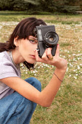 Side view of focused young female in casual clothes taking photo on vintage camera while sitting on grassy lawn with flowers - ADSF49593