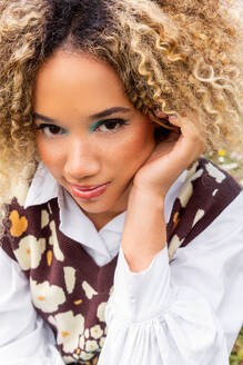 Smiling ethnic young woman with curly blonde hair looking at camera while resting her face on her hand and wearing a white collared shirt beneath a patterned sweater vest adorned with flower shapes - ADSF49595