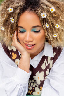 High angle of ethnic young woman with curly blonde hair looks down while resting her chin on her hand and wearing a white collared shirt beneath a patterned sweater vest adorned with flower shapes - ADSF49597