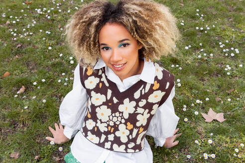 High angle of smiling ethnic young woman with curly blonde hair looking at camera while sitting on garden and wearing a white collared shirt beneath a patterned sweater vest adorned with flower shapes - ADSF49598