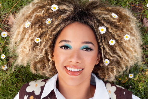 Top view of young ethnic woman with curly hair decorated with delicate white daisies looking at camera while lying on garden in daylight - ADSF49600