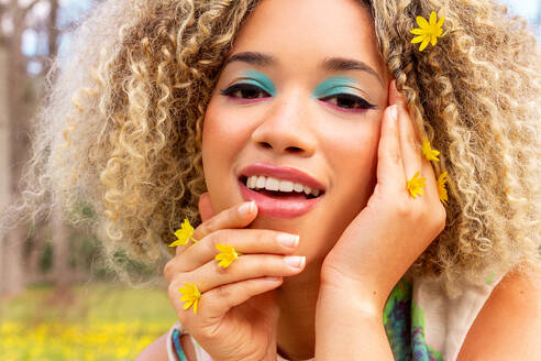 Close-up portrait of a radiant young ethnic woman with curly golden hair, adorned with small yellow flowers and make up looking at camera and smiling - ADSF49603