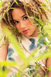 Closeup of ethnic young woman with curly blonde hair stands amidst tall green reeds while looking at camera - ADSF49606