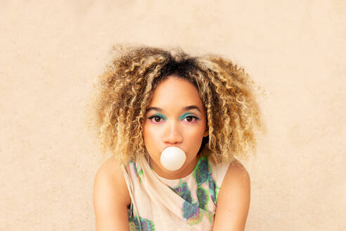 Close-up of ethnic young woman with curly blonde hair blowing a bubble with a pink bubblegum while looking at camera against a textured beige background - ADSF49608