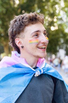 Young man with a joyful expression, draped in a pastel flag, with rainbow-colored stripes painted on his cheeks, symbolizing LGBTQ+ pride amidst a natural setting - ADSF49609