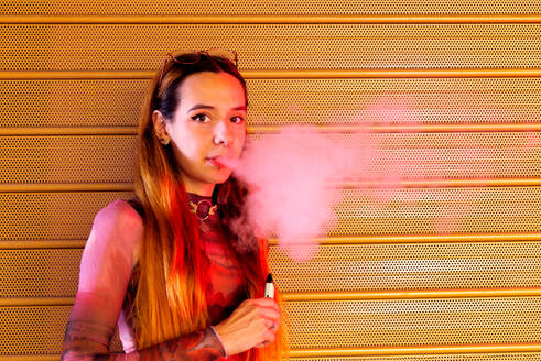 Seitenansicht einer jungen Frau mit roten Haaren, die vor einem orangefarbenen perforierten Hintergrund steht, eine E-Zigarette hält und eine Dampfwolke ausatmet, während sie in die Kamera schaut - ADSF49612