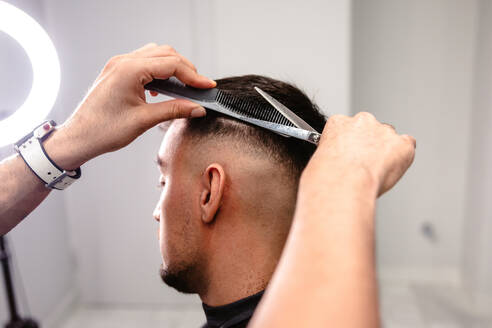 Close-up of a barber's hands combing and cutting the hair on the back of a man's head in a white-walled salon with circular light. - ADSF49617