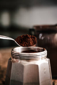 Close-up of spoon ground coffee from a jar to a stainless steel stovetop espresso maker set on a speckled countertop - ADSF49618
