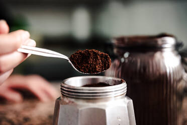 Close-up of anonymous person hand transferring freshly ground coffee from a jar to a stainless steel stovetop espresso maker set on a speckled countertop - ADSF49619