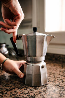 Anonymous young individual hands expertly assembling a stainless steel stovetop espresso maker on a speckled countertop with ambient light filtering through a nearby window - ADSF49620