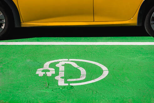 White symbol of electric car charger with plug placed on green ground near yellow car in city street - ADSF49621