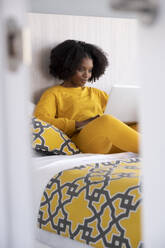 Young African American woman in yellow attire sitting on a bed with patterned cushions engrossed in her laptop in a well lit room - ADSF49626