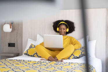 Cheerful black woman in a yellow outfit enjoys content on her laptop while wearing headphones seated on a bed with a stylish design - ADSF49627