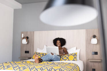 Cheerful black woman with curly hair lounges on a modern bed engrossed in her laptop surrounded by contemporary room decor - ADSF49628