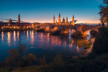 Arched stone bridge and illuminated Basilica of Nuestra Senora del Pilar over Ebro river against clear blue sky during dusk - ADSF49631