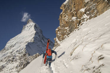 Back view of unrecognizable person with ski equipment walking on snow covered mountain slope during sunny day at Swiss Alps - ADSF49647