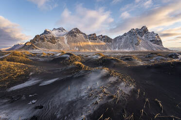 Majestic Stokksnes mountains in Islandia stand tall against a painted sky their snowy peaks contrasting with the dark patterned sands below the golden sunlight softly illuminates the landscape - ADSF49673