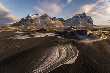 Majestic Stokksnes mountains in Islandia stand tall against a painted sky their snowy peaks contrasting with the dark patterned sands below the golden sunlight softly illuminates the landscape - ADSF49674