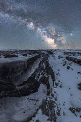 Weitläufige Luftaufnahme einer schneebedeckten Landschaft, die von einem tosenden Wasserfall durchschnitten wird, mit einer sternenübersäten Milchstraße, die den dunklen Himmel über Island erhellt - ADSF49679