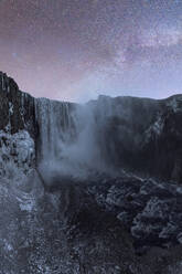 Majestic view of a powerful waterfall descending amidst rugged cliffs, enveloped by mist, under a dim, twilight sky with hints of pink and purple hues in Iceland - ADSF49680